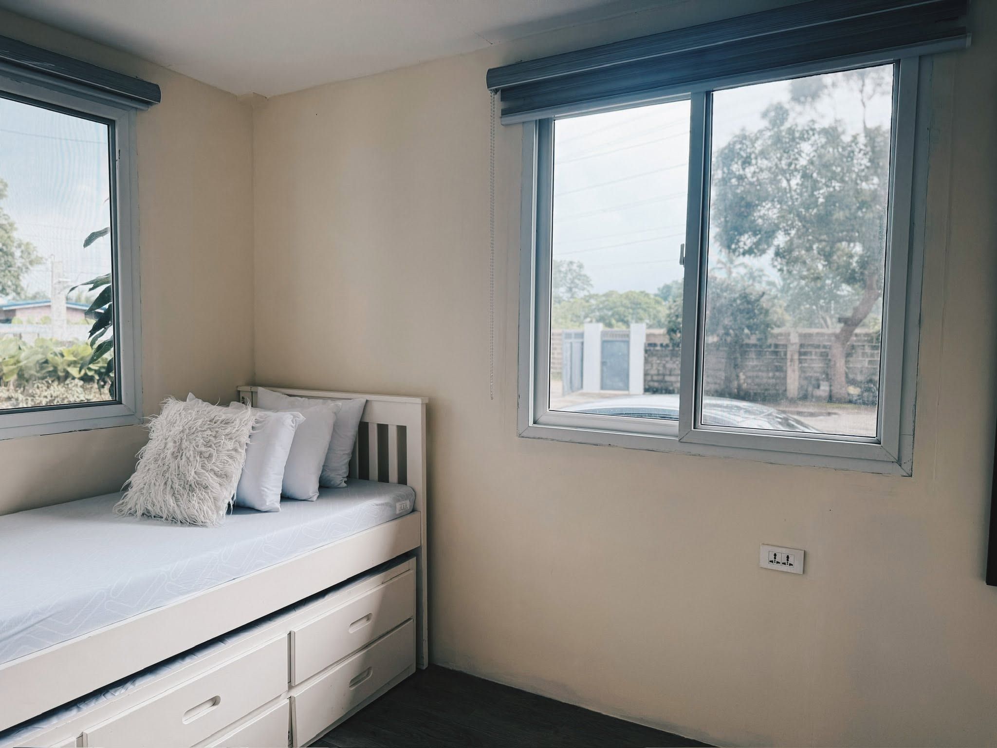 White daybed with trundle drawers and pillows, beside two windows looking out to trees