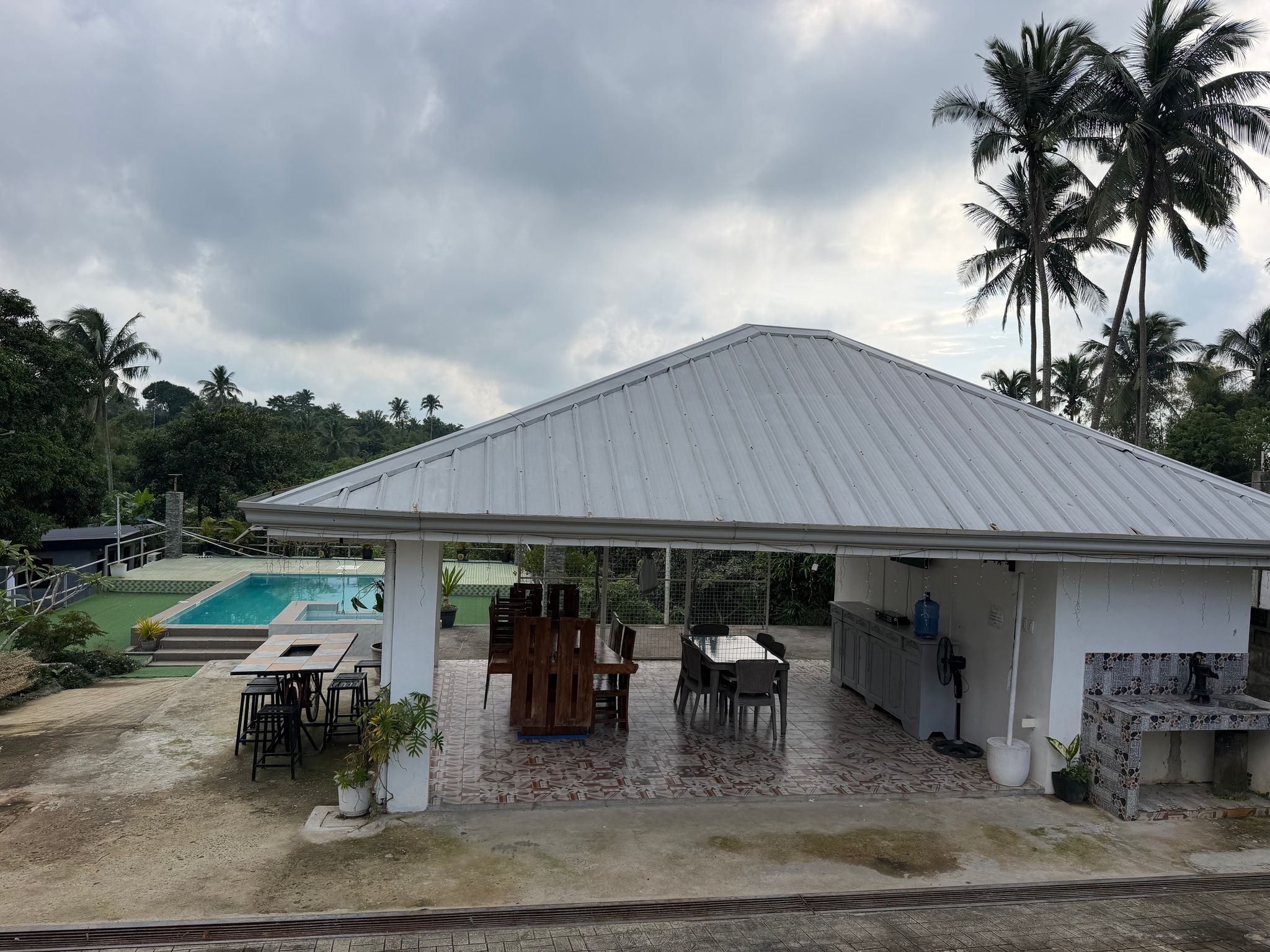The open-sided pavilion with long wooden dining tables, a high table with stools and a sink counter, the pool beyond