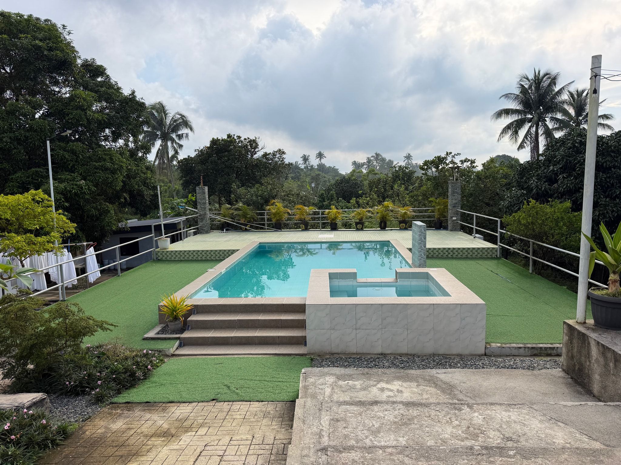 Steps up to the pool deck, with the kiddie pool in front, potted palms along the railing and trees beyond