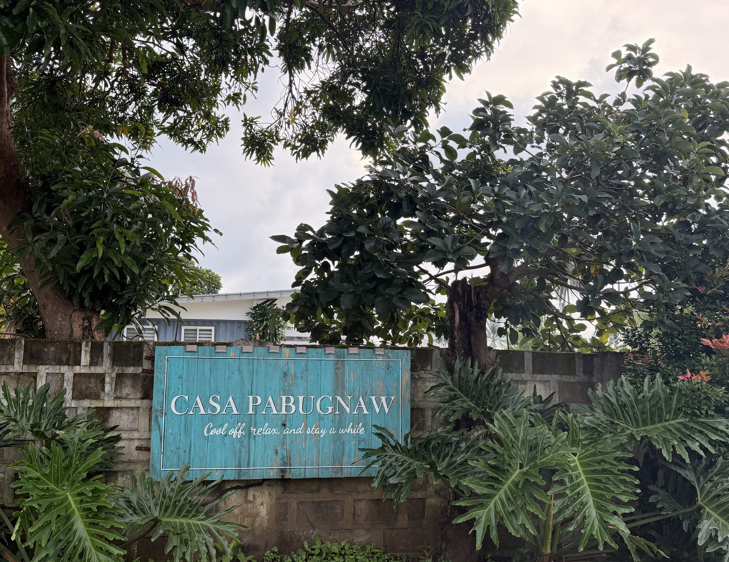 The teal Casa Pabugnaw sign on the front wall, reading “Cool off, relax, and stay a while”, among monstera and shade trees