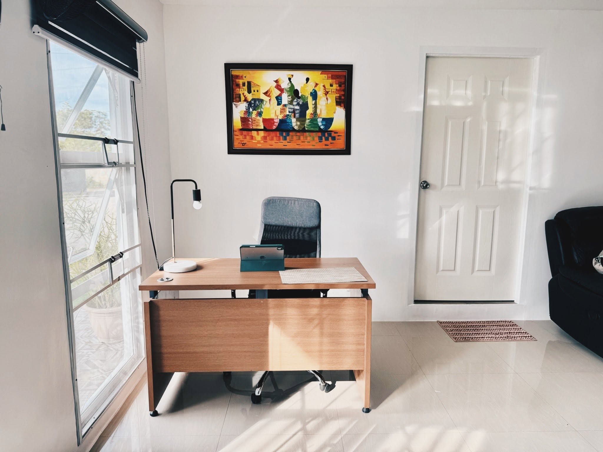 Wooden work desk with a lamp and an office chair beside a tall window, under a colourful painting
